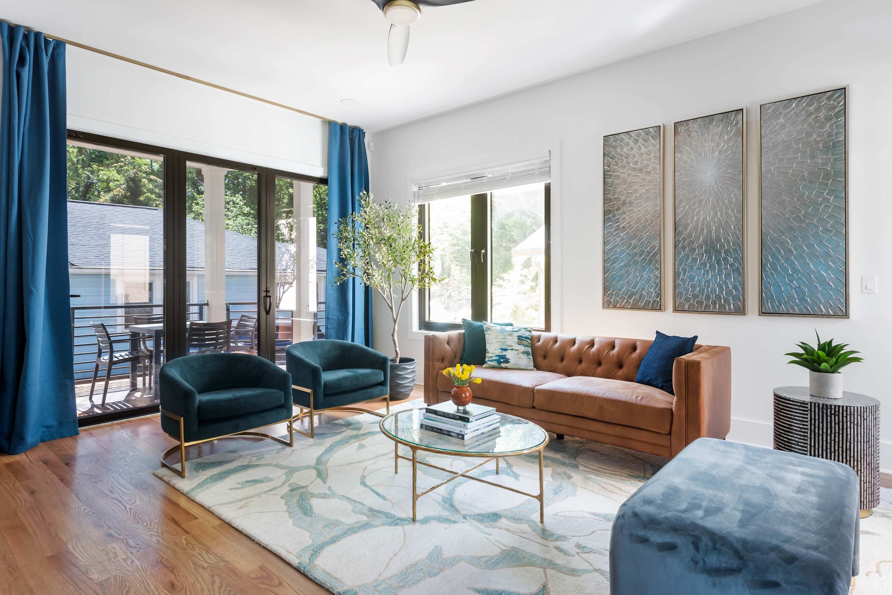 Un salon moderne avec un canapé en cuir, deux fauteuils, une table basse en verre et une grande fenêtre ornée de rideaux bleus.