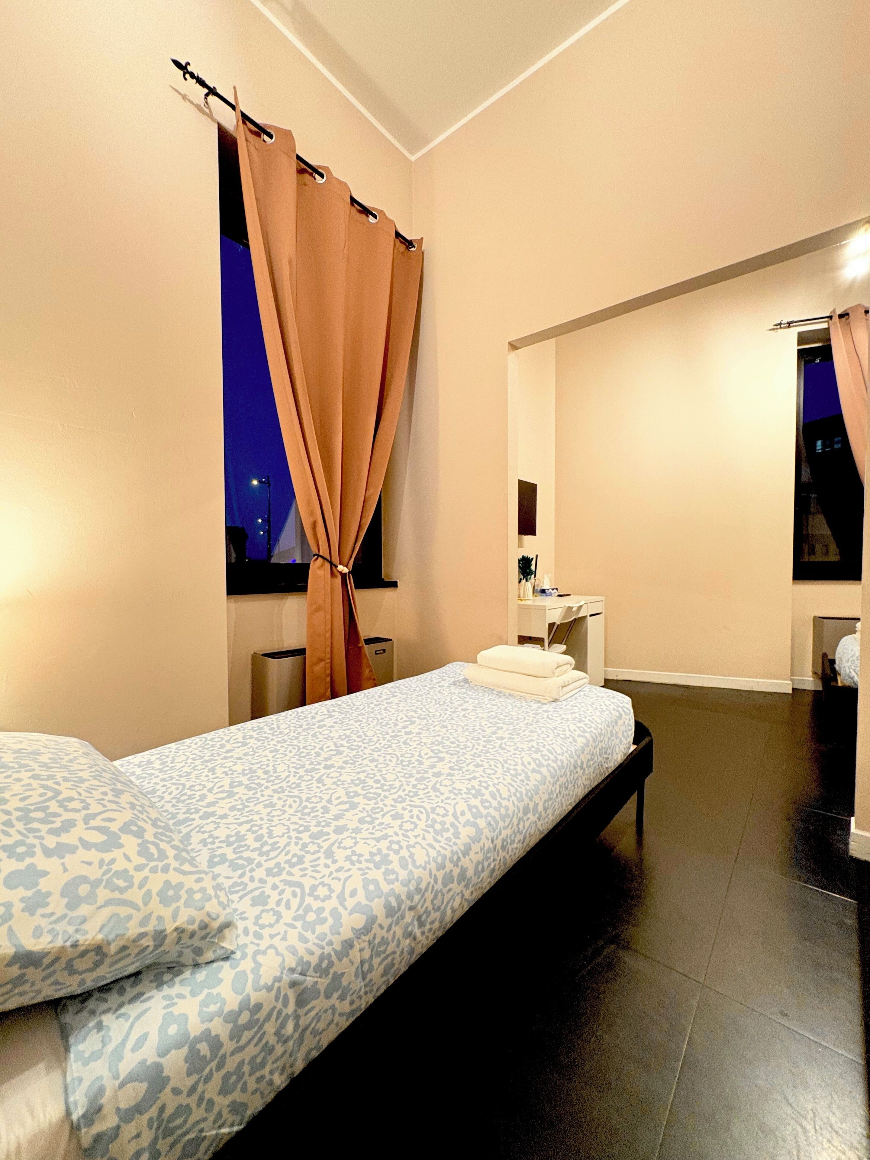 Une chambre d’hôtel moderne, équipée d’un lit, d’un petit bureau, d’un miroir et d’une salle de bain visible à travers une porte ouverte.