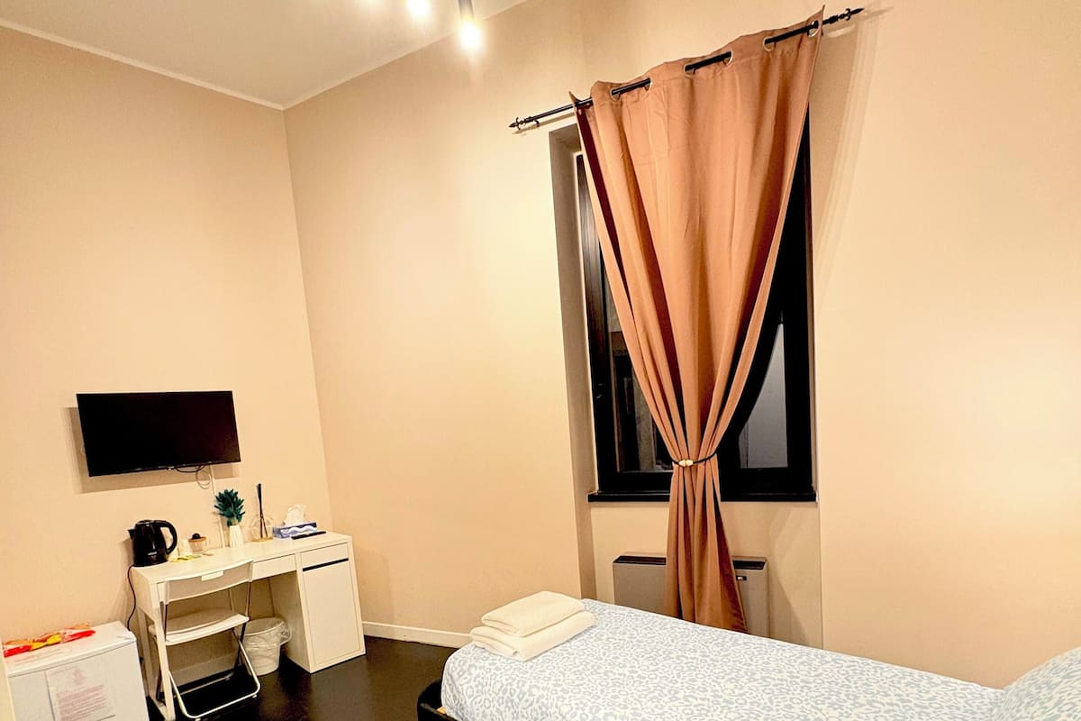 A bedroom with a bed, a desk, a chair, a TV, and a window with curtains.