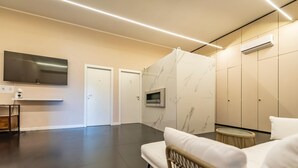 Une pièce moderne et minimaliste, dotée d’un plancher foncé, d’un canapé blanc, d’une petite table ronde, d’un téléviseur fixé au mur et d’un climatiseur mural.