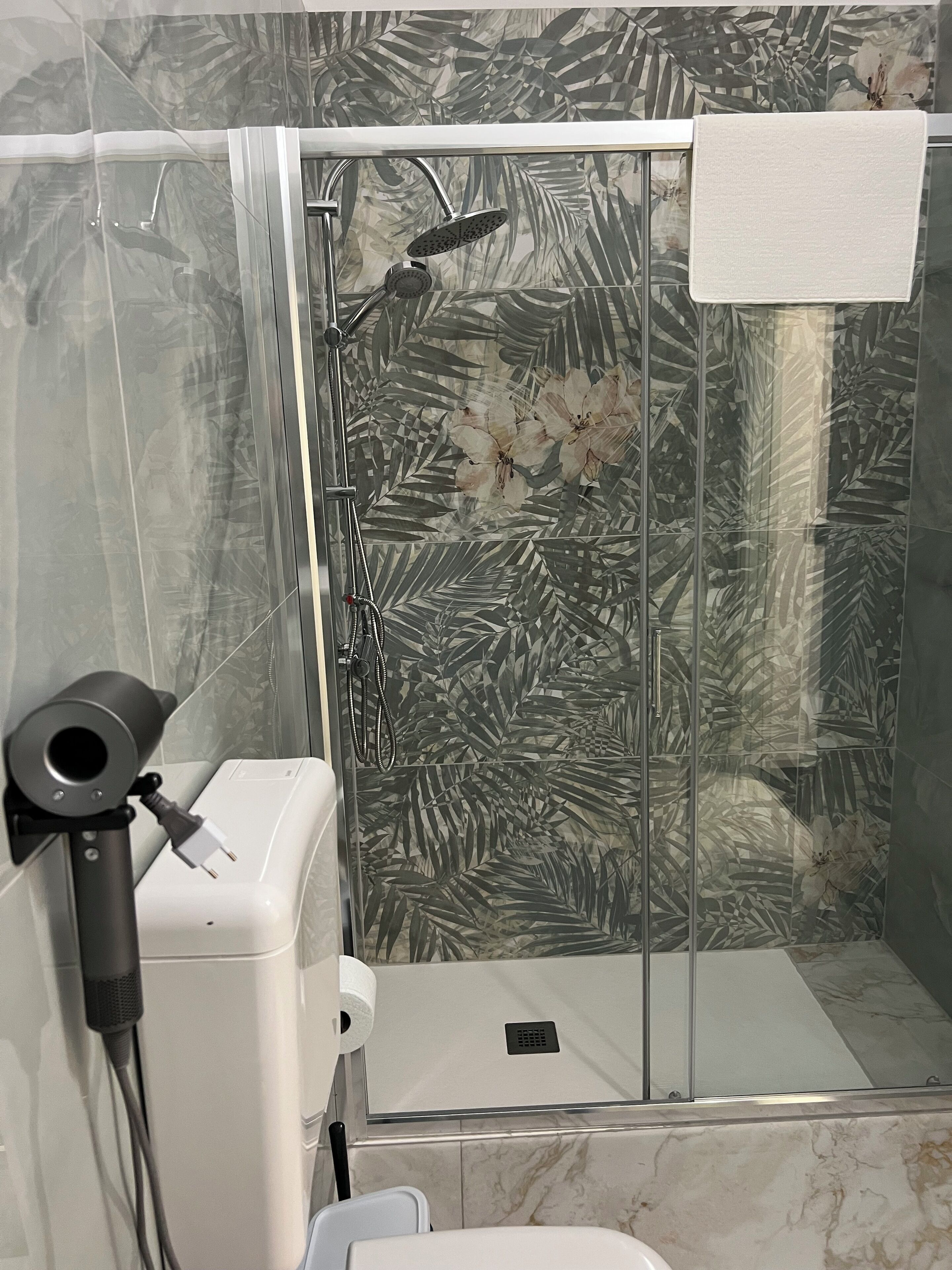 Una ducha con paredes de vidrio, un diseño de azulejos con motivos florales, una toalla colgada en la puerta y un secador de pelo conectado.