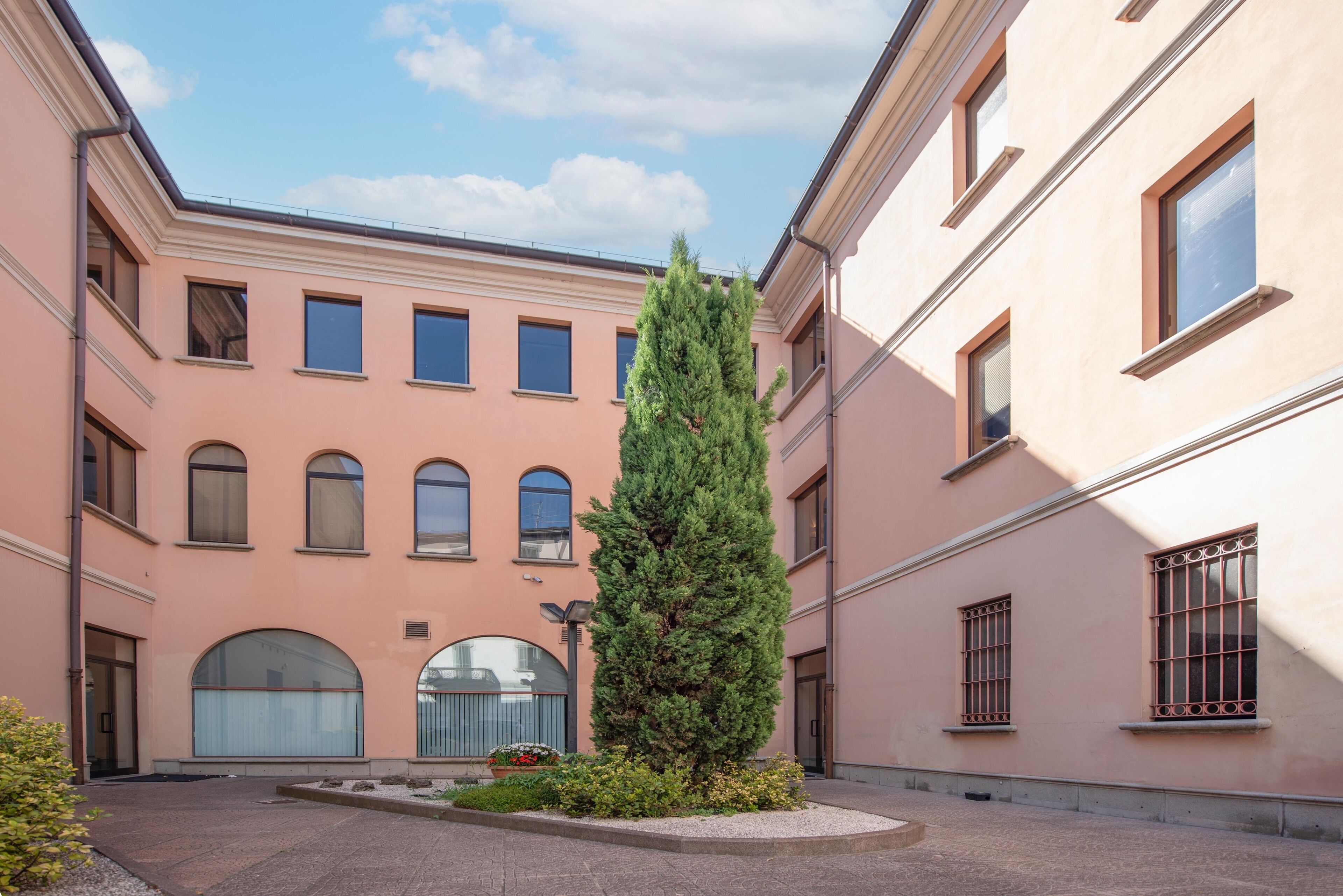 Un patio con un alto árbol de hoja perenne, un edificio con ventanas arqueadas y un camino pavimentado.