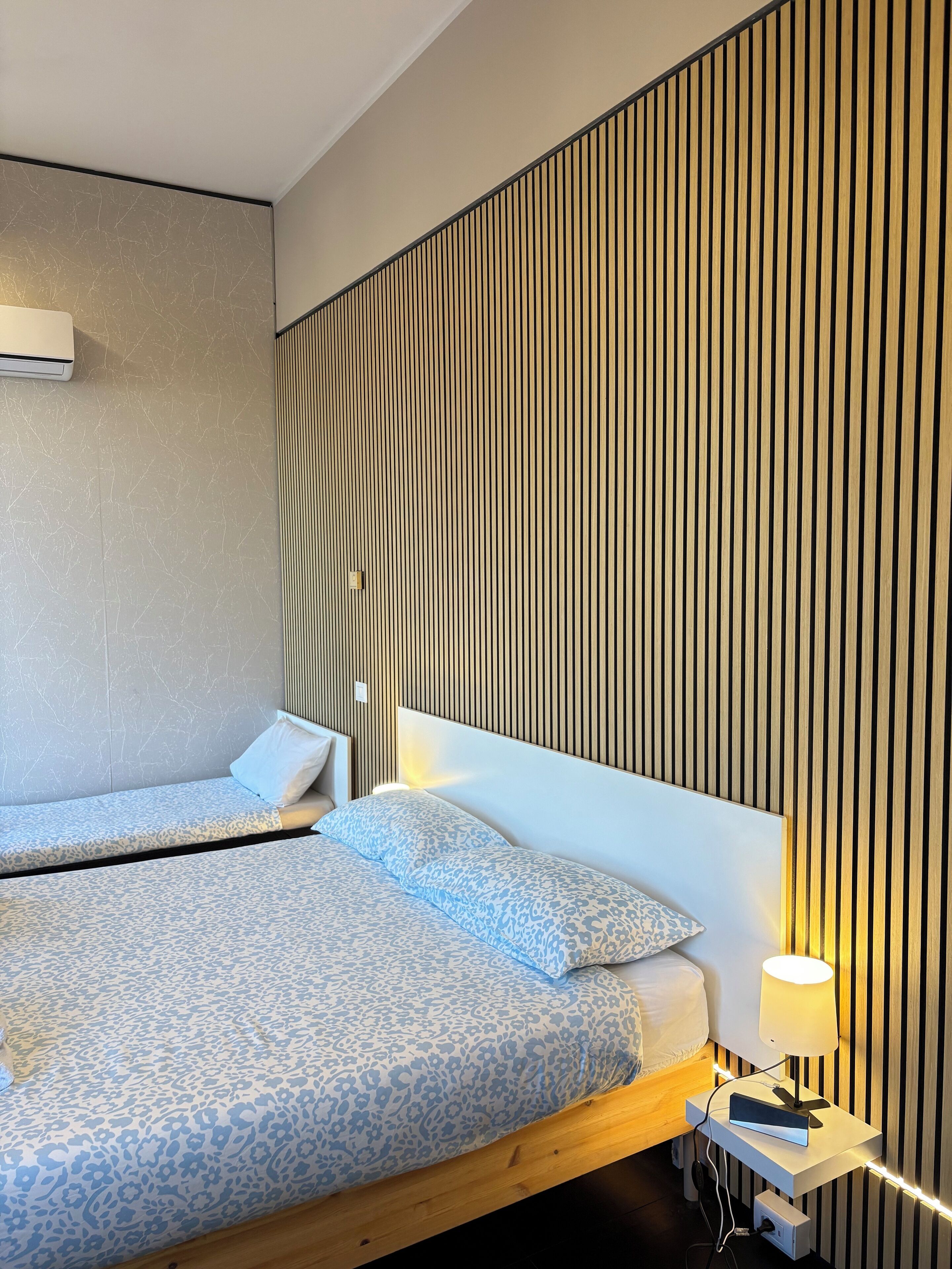 Une chambre d’hôtel moderne avec deux lits, une tête de lit en bois, une table de chevet avec une lampe et un climatiseur fixé au mur.