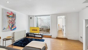 A modern living room with a sofa, a coffee table, and a TV on a stand. There is a large window with a view of a garden. - Group & Contractor-Friendly 4Bed House (Brighton and Hove)
