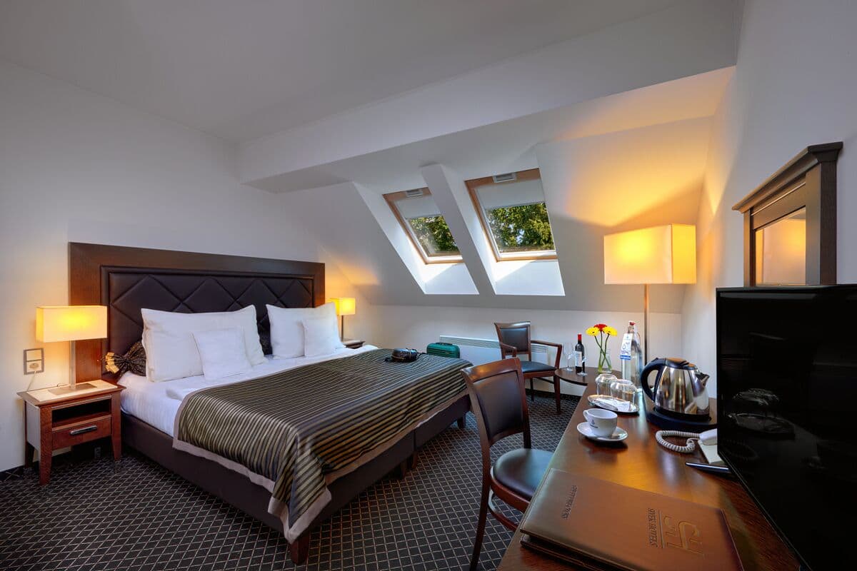 A hotel room with a bed, a desk with a laptop, a chair, a TV, and a window with a view of trees.