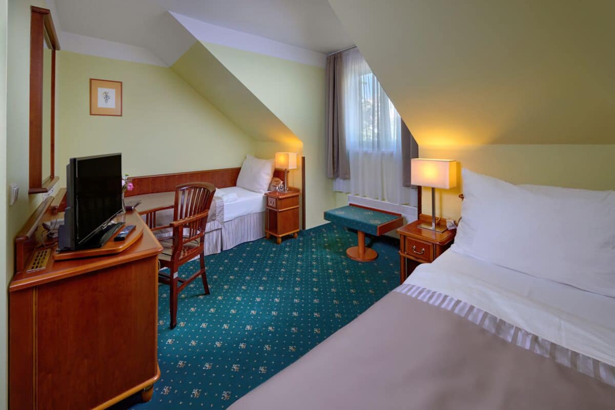 A hotel room with a bed, a desk with a computer, a chair, a small table, and a window with curtains.