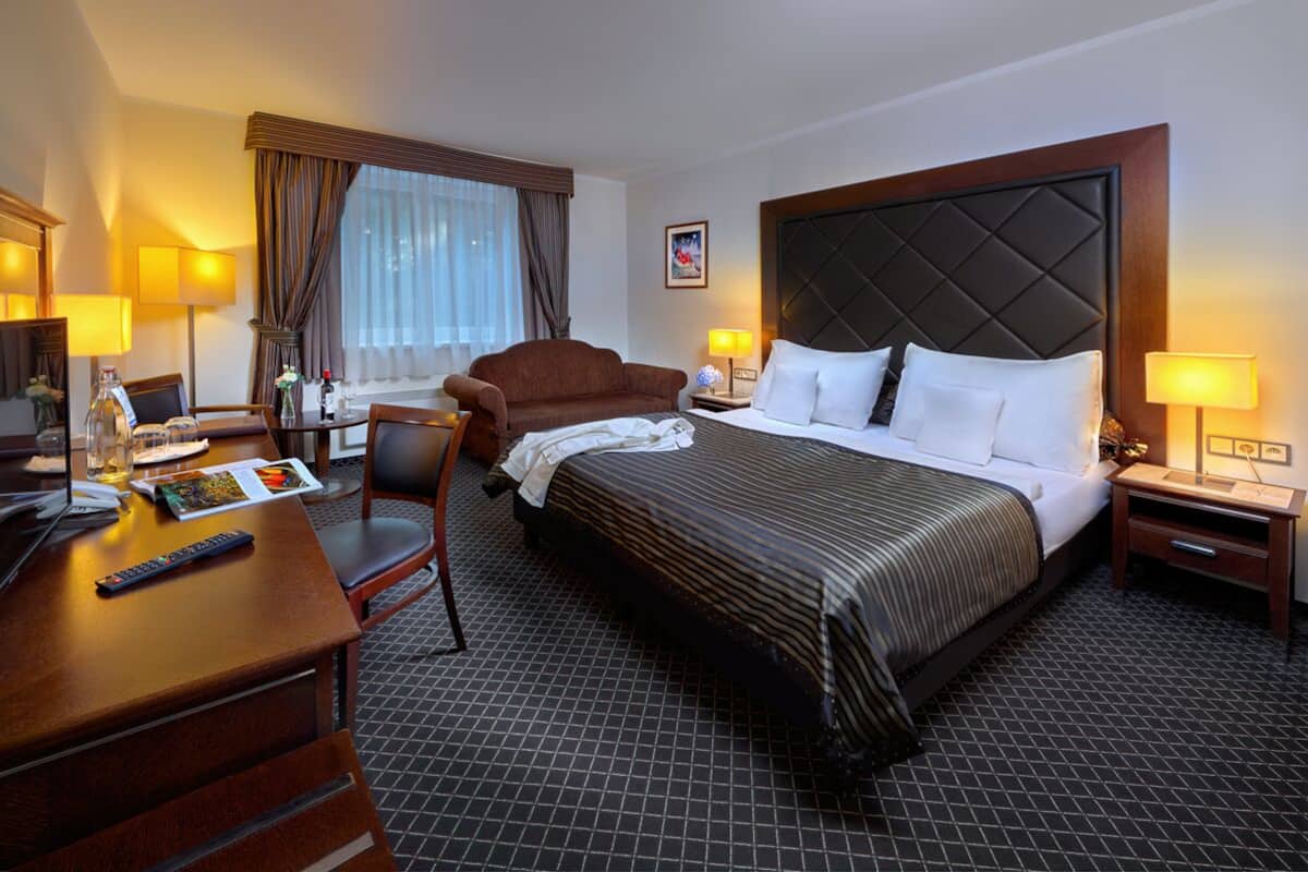 A hotel room with a large bed, a desk with a computer and remote control, a chair, a small table with a lamp, and a window with curtains.