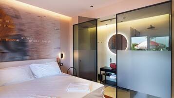 A modern hotel room with a large bed, a glass partition, a round mirror, and a view of greenery outside.