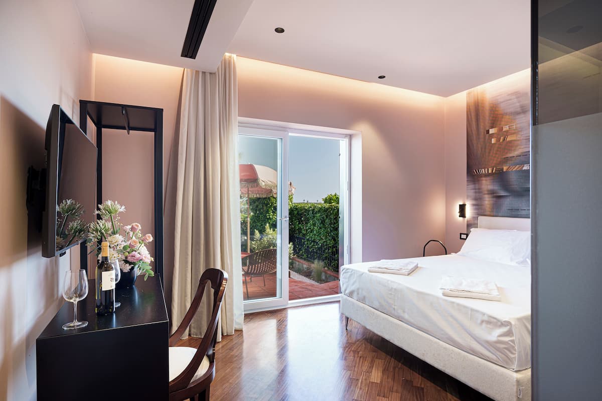 A hotel room with a bed, a desk with a wine bottle, a chair, and a large window with a view of greenery.
