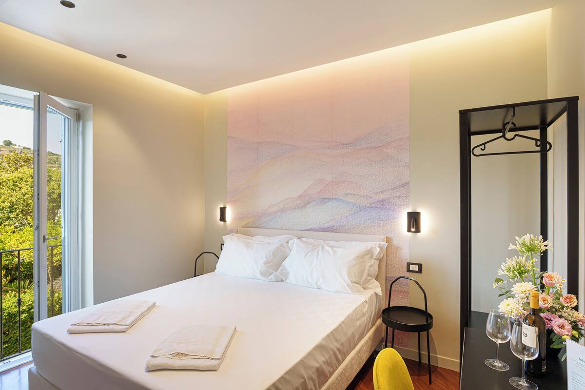 A modern hotel room with a large bed, a view of greenery through a window, a wall-mounted light, and a table with a bottle and glasses.