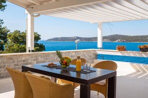 Un espacio de comedor al aire libre cubierto con una mesa, sillas de mimbre, un frutero, una botella y vasos. Se ve una piscina y vistas al mar y las colinas al fondo.