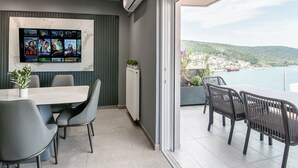 Una habitación moderna con un televisor grande mostrando carteles de películas, un rincón comedor con una mesa y sillas, y un balcón con vistas a un pueblo costero.