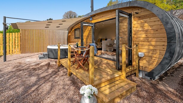 A modern, cylindrical wooden cabin with a deck and hot tub, surrounded by a wooden fence and gravel.