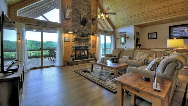A spacious living room with a fireplace, wooden ceiling, and large windows offering a view of the outdoors. - Pet-friendly, family-sized luxury cabin rental, hot tub, WiFi, firepit (Mineral Bluff)