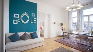 A modern living room with a white sofa, a teal accent wall, and a dining area with a table and chairs. - Waterfront Apt w/ River Views & Trendy Location (Praha 5)