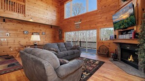 A cozy cabin interior with a fireplace, large windows, and a television.