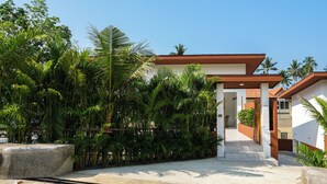 Une maison moderne, à l’extérieur blanc et au toit brun, entourée d’une végétation luxuriante et de palmiers.