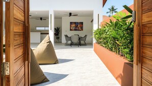 Une porte en bois ouverte donne sur une cuisine et un coin repas modernes, avec un ventilateur de plafond, une table à manger avec des chaises et une grande fenêtre.