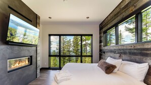 A modern bedroom with a large bed, a fireplace, and a TV mounted on the wall. - Ski Access, Overlooking the Lake + Private Hot Tub & Pets Considered (Whitefish)
