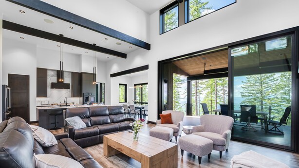 A modern living room with a sofa, coffee table, and chairs, a kitchen area, and large windows with a view of trees. - Ski Access, Overlooking the Lake + Private Hot Tub & Pets Considered (Whitefish)