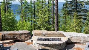 A stone fire pit with seating, surrounded by large rocks, overlooking a forested area and a lake. - Ski Access, Overlooking the Lake + Private Hot Tub & Pets Considered (Whitefish)
