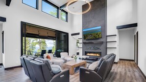 A modern living room with a fireplace, leather sofas, and a large window overlooking a scenic landscape. - Ski Access, Overlooking the Lake + Private Hot Tub & Pets Considered (Whitefish)