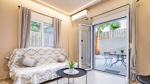 A modern living room with a sofa, a small round table with a plant, and a sliding glass door leading to an outdoor patio. - Dom Azure Elegant Studios (Chania)