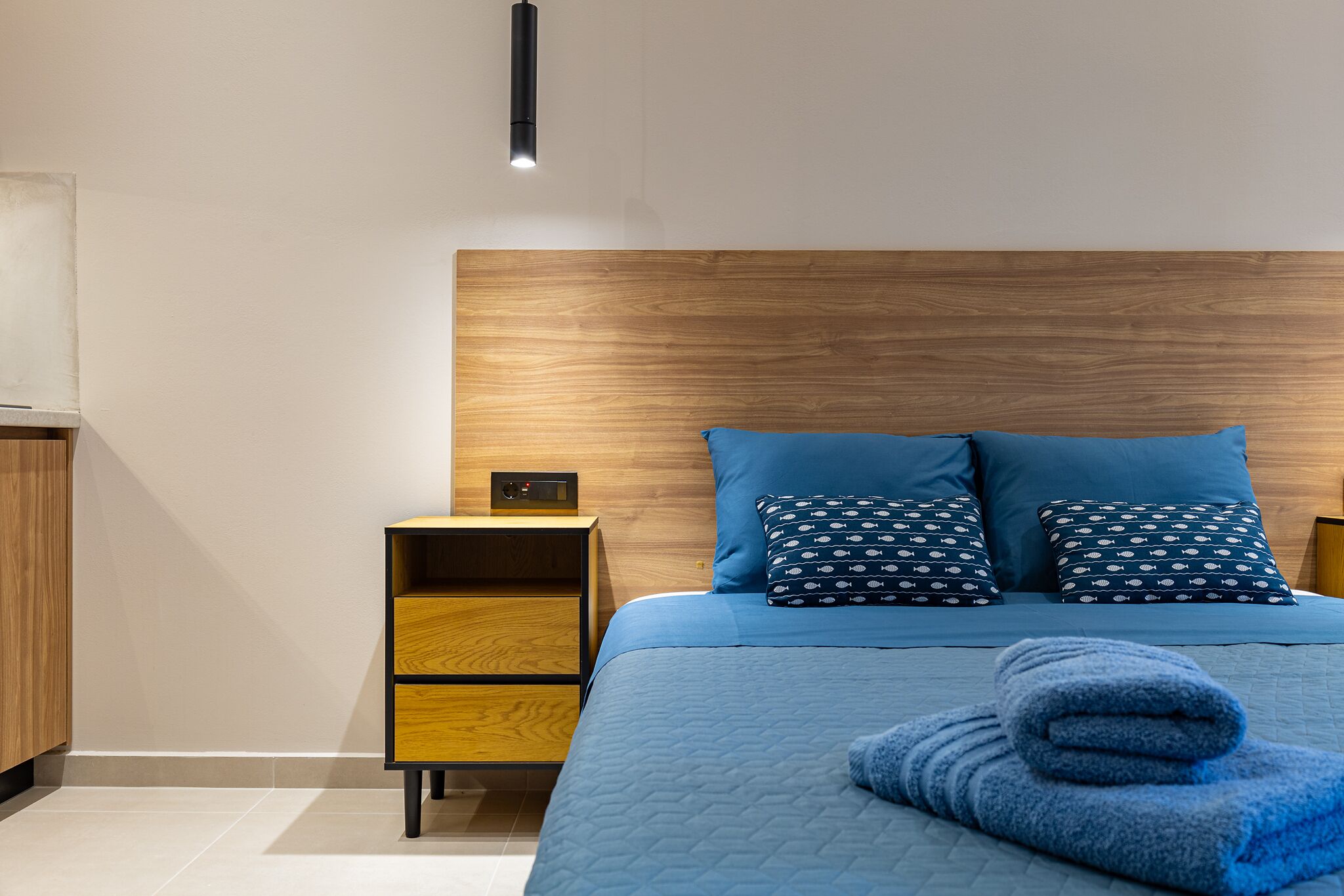 A modern bedroom with a wooden headboard, a blue bedspread, matching pillows, a nightstand with a lamp, and a neatly folded towel on the bed.