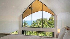 A bedroom with a large window overlooking a tropical landscape and the sea.