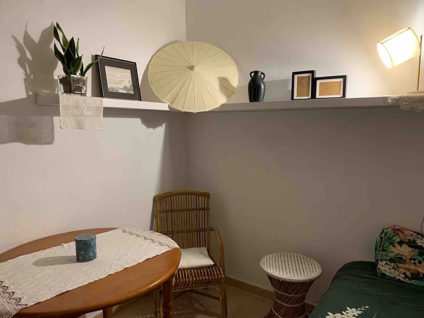 Una habitación con una mesa redonda, una silla de mimbre, un taburete pequeño, una cama con colcha estampada, un estante con una planta, un jarrón y cuadros enmarcados.