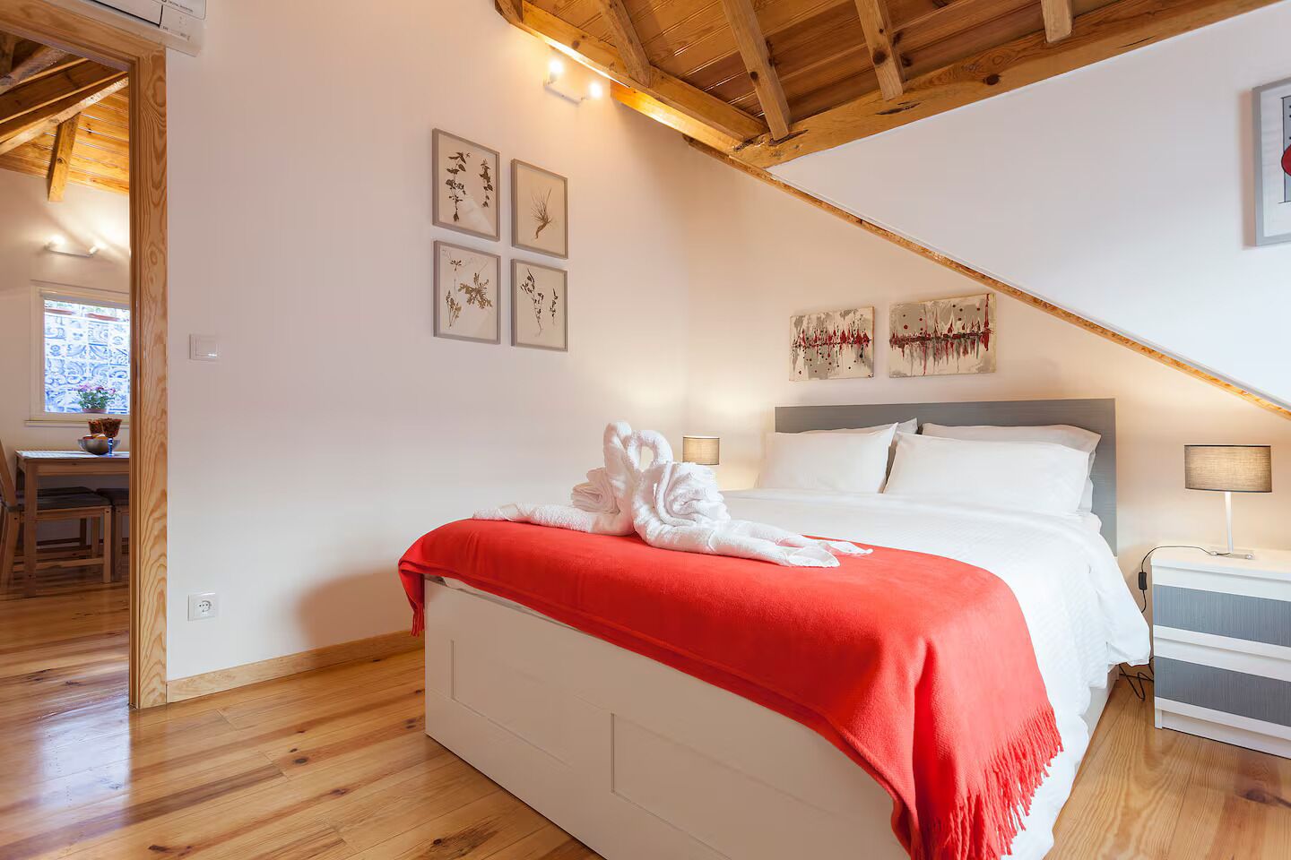 A bedroom with a bed, a red blanket, and a white towel. There are wall art pieces and a window with a view.