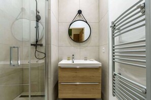 A bathroom with a glass shower enclosure, a round mirror, a white sink, and a towel rack.