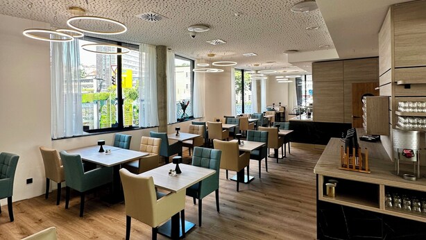 A modern restaurant with a wooden floor, round tables, and chairs. There are large windows with views of buildings outside.