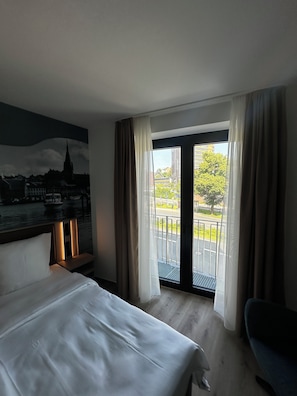 A hotel room with a bed, a chair, a painting of a cityscape, and a view of a building and greenery through the window. - Central Hotel (Flensburg)