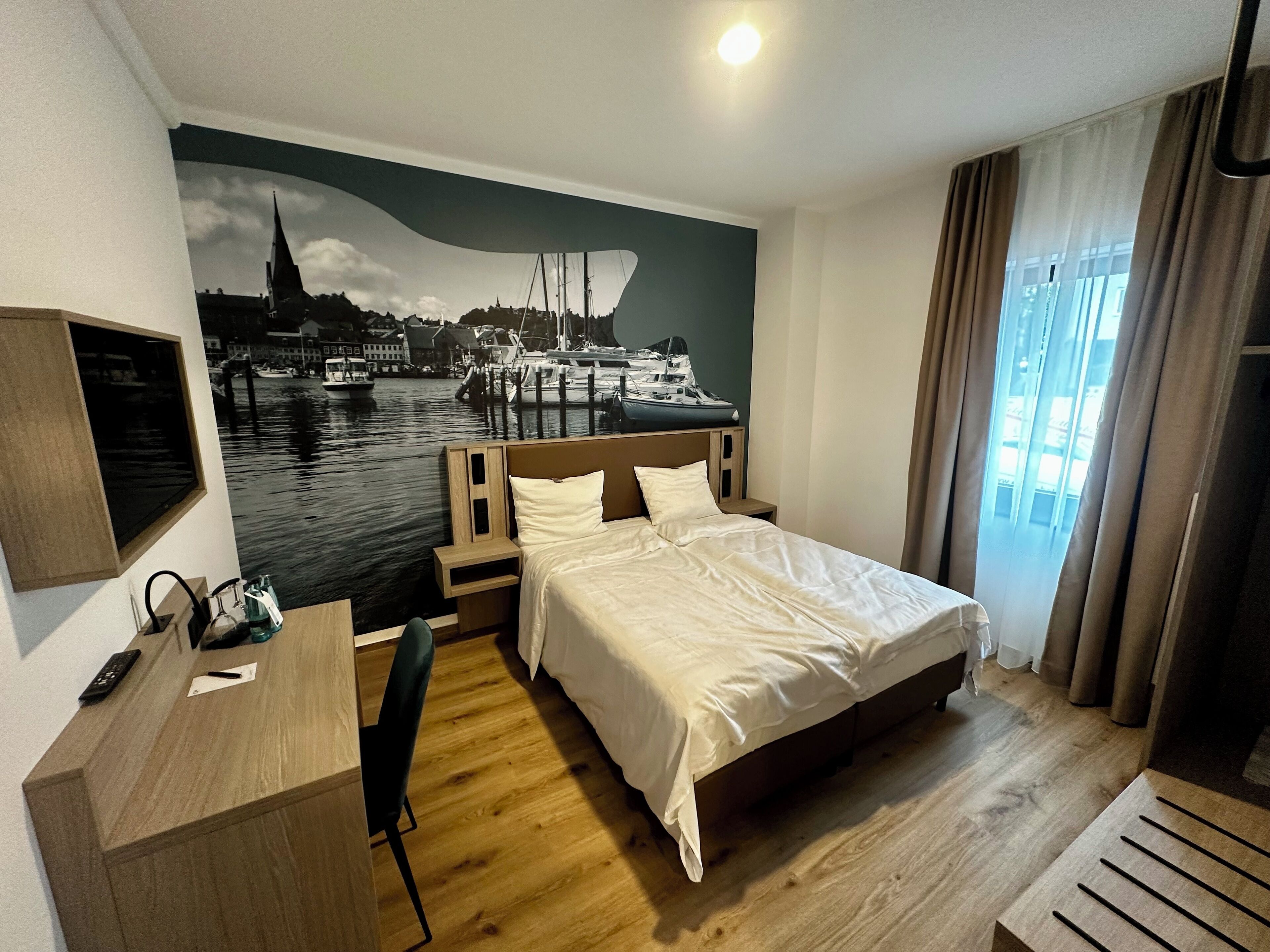 A hotel room with a bed, a desk, a chair, a nightstand, and a wall mural of a harbor scene.