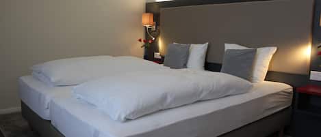 A double bed with white linens and two pillows, a nightstand with a lamp, and a wall-mounted light fixture.