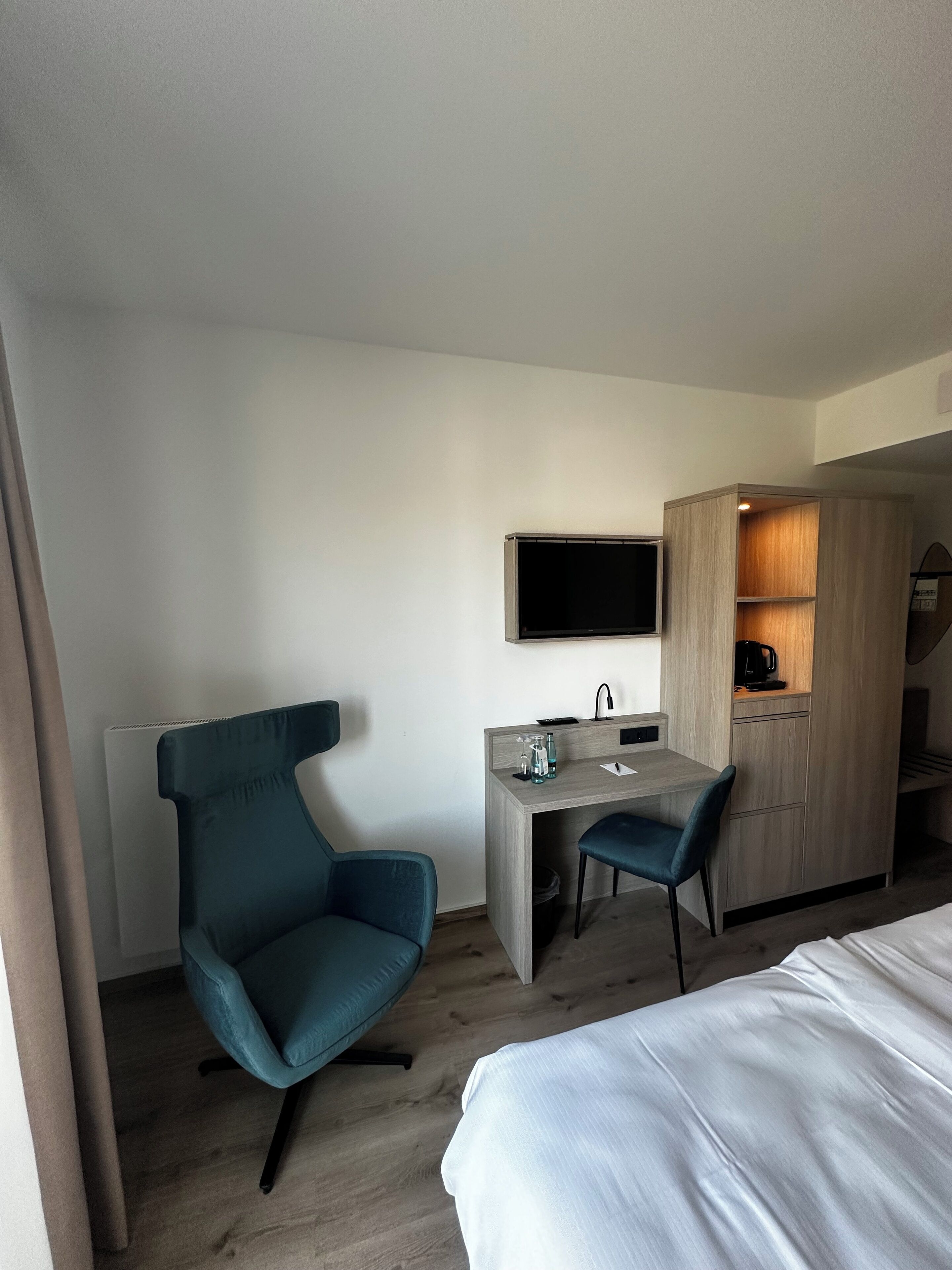 A modern hotel room with a bed, a desk with a chair, a TV mounted on the wall, and a wardrobe.