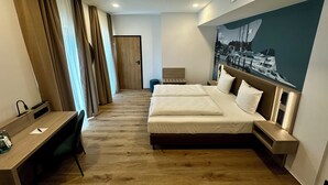 A hotel room with a large bed, a desk, and a chair. There is a door leading to another room and a wall with a picture of boats.