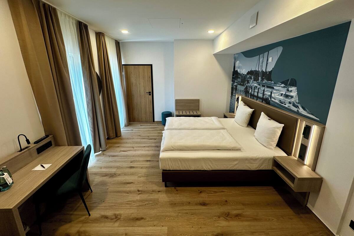 A hotel room with a large bed, a desk, and a chair. There is a door leading to another room and a wall with a picture of boats.