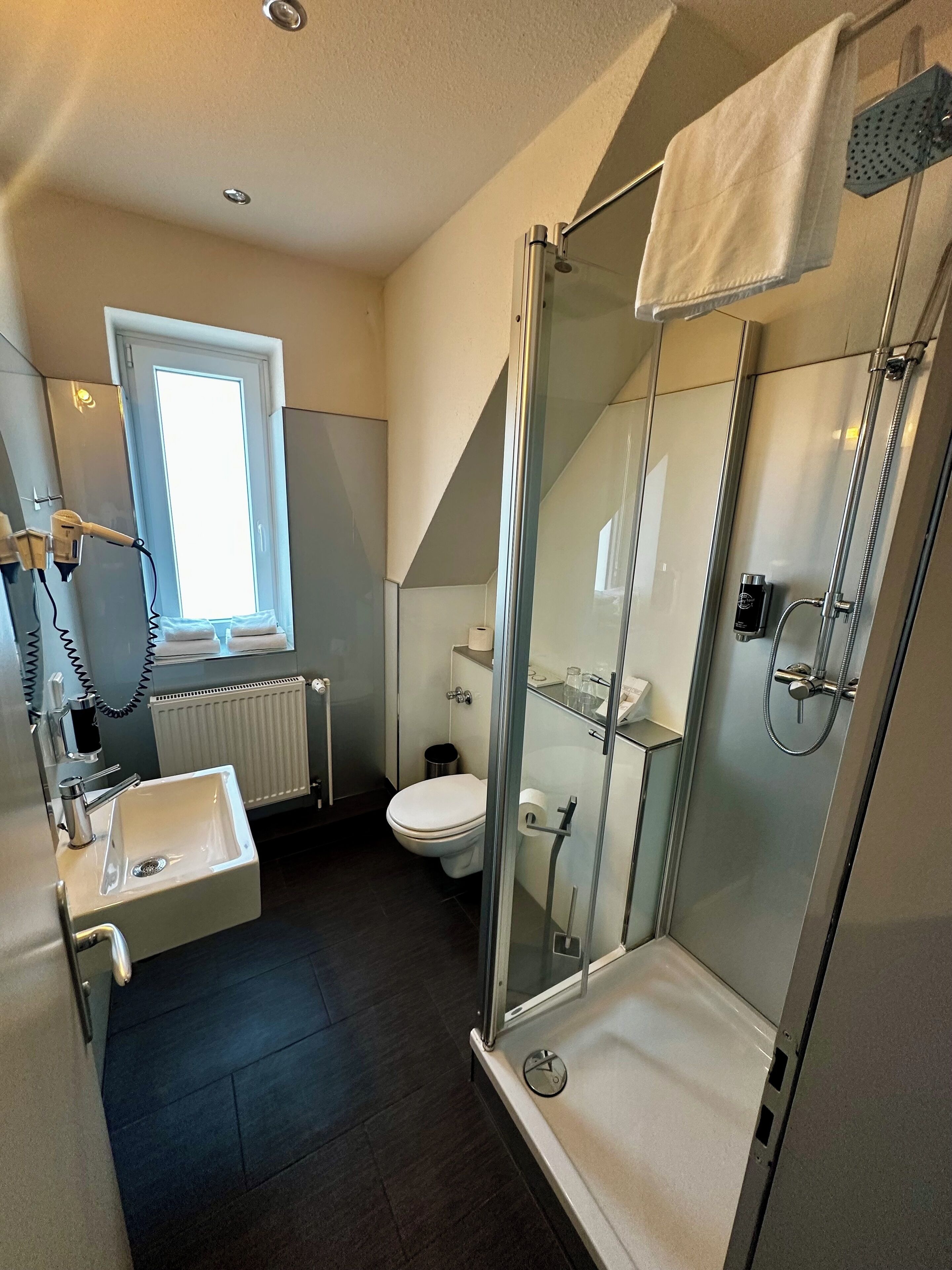 A modern bathroom with a glass shower enclosure, a sink with a mirror, a toilet, and a window providing natural light.