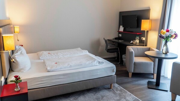 Habitación de hotel con cama, escritorio con silla, televisor y una pequeña mesa con un jarrón de flores.