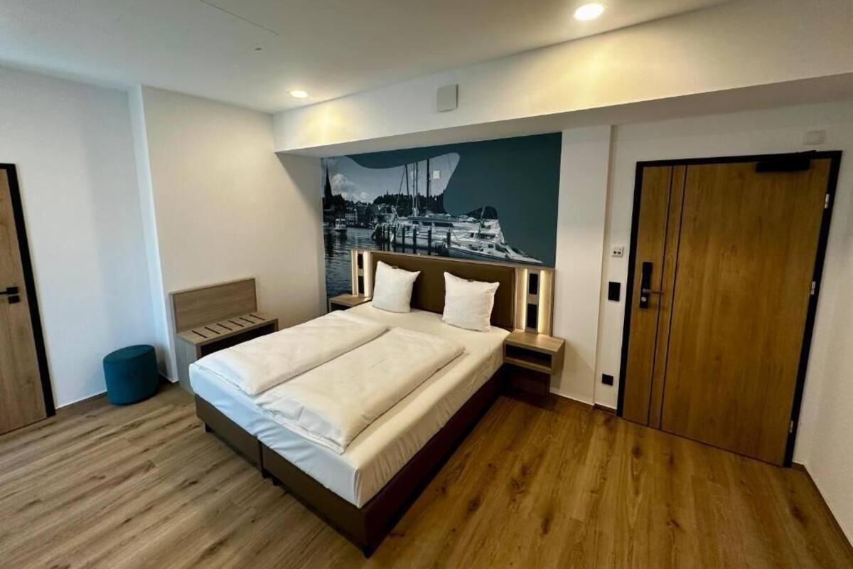 A modern bedroom with a large bed, wooden flooring, a wall-mounted headboard, and a door.