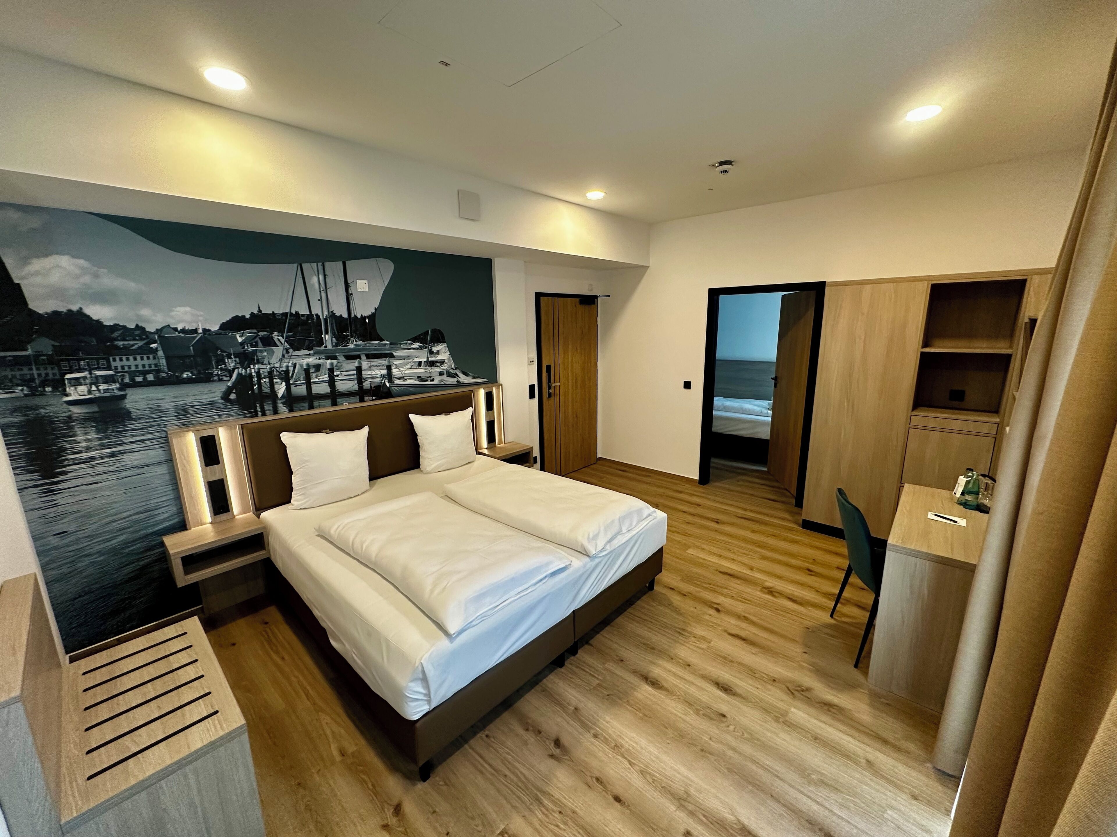 A modern hotel room with a large bed, wooden flooring, a desk, and a view of a marina through a mural.