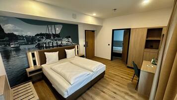 A modern hotel room with a large bed, wooden flooring, a desk, and a view of a marina through a mural.
