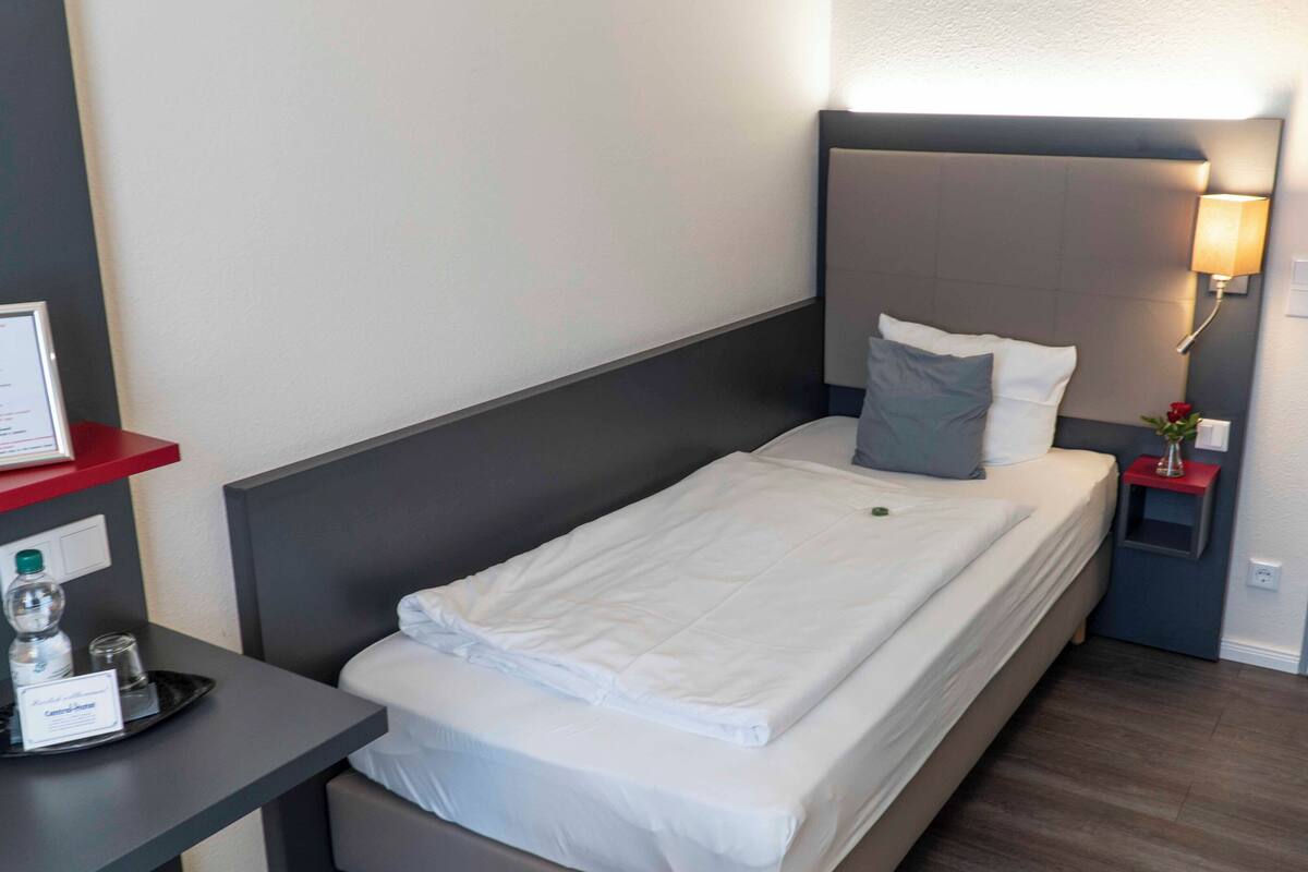A neatly made bed with white linens, a bedside table with a lamp, and a small nightstand with a vase of flowers.