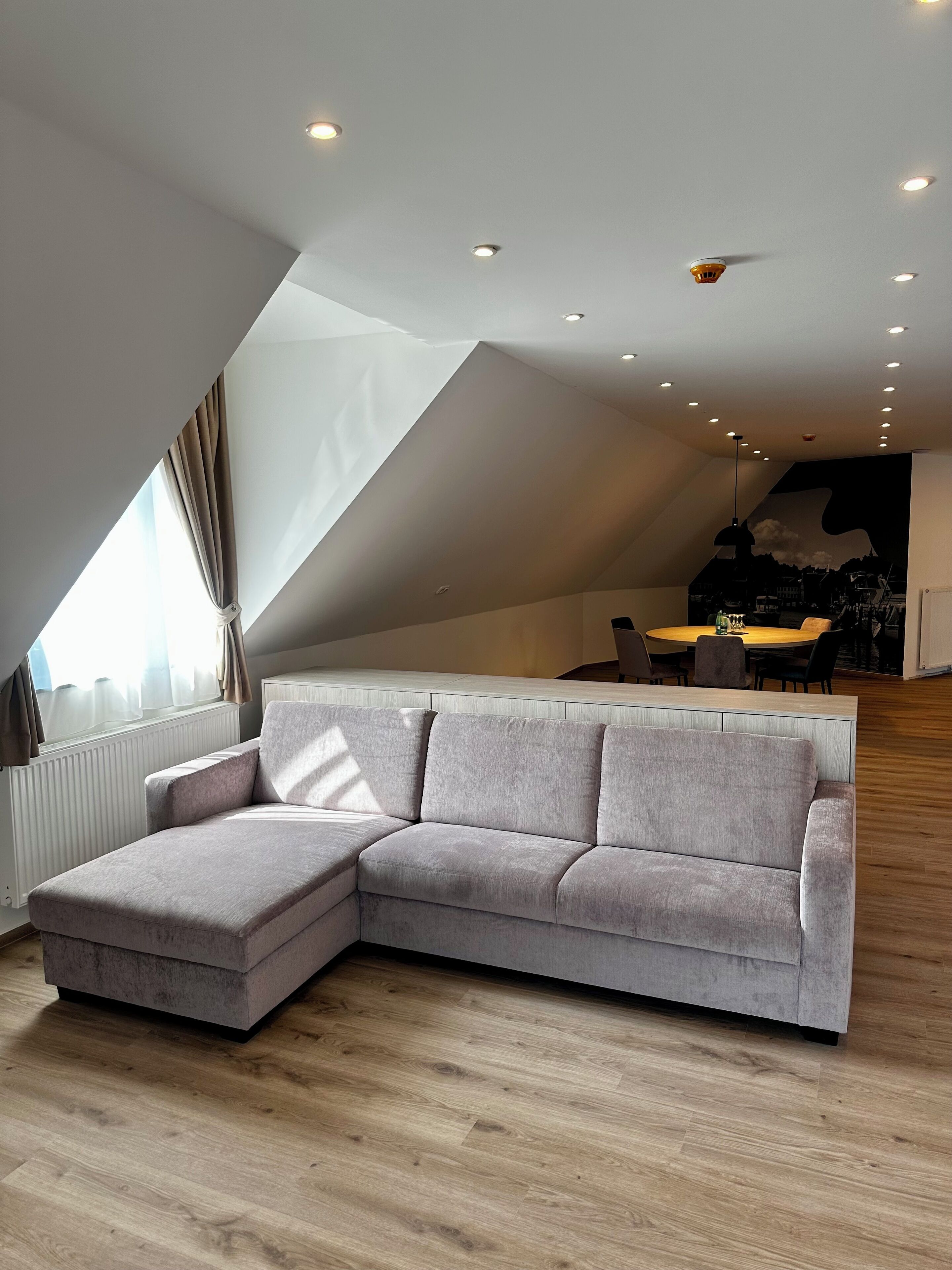 Ein modernes Wohnzimmer mit einem grauen Ecksofa, einem Essbereich mit rundem Tisch und einer schrägen Decke mit eingelassener Beleuchtung.