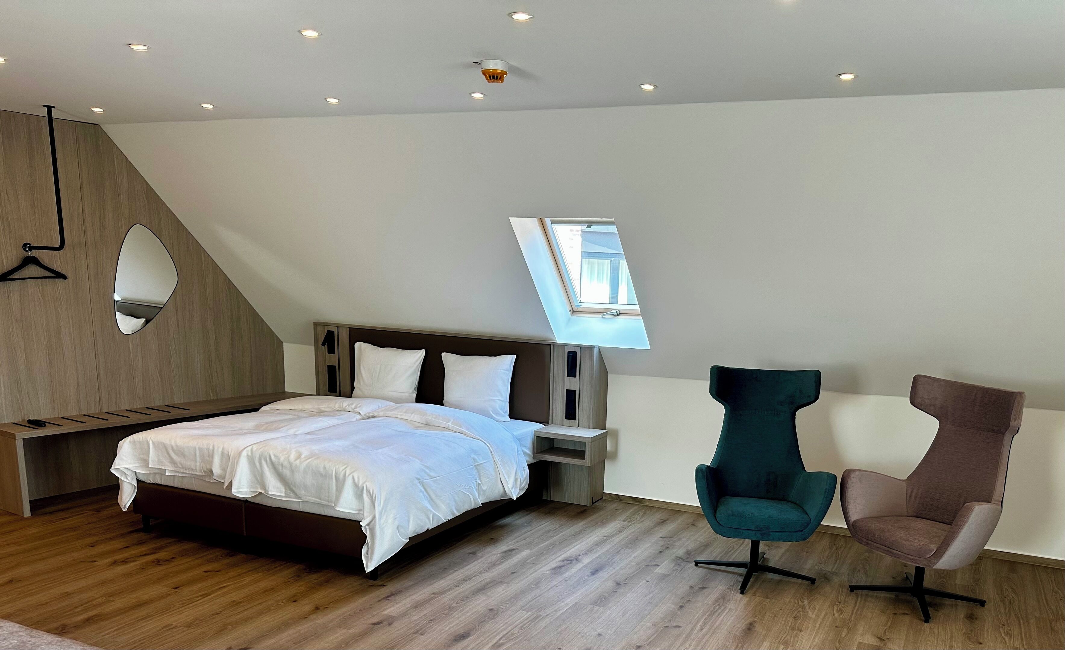 Ein modernes Schlafzimmer mit schrägen Wänden, einem Oberlicht, einem Bett, zwei Stühlen und einer hölzernen Sitzbank.