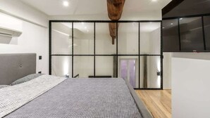 A modern bedroom with a large bed, a wooden ceiling beam, and a sliding glass door leading to a bathroom.