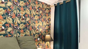 Une chambre à coucher avec un papier peint à motifs floraux, un lit avec une literie verte et violette, une lampe et un rideau bleu.