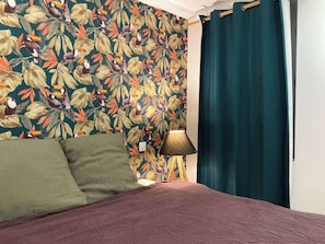 A bedroom with a floral wallpaper, a bed with green and purple bedding, a lamp, and a blue curtain.